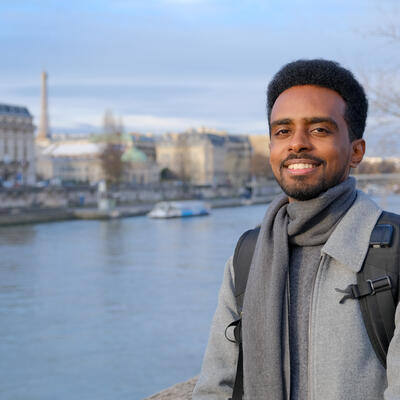 Person standing by a river in a city, wearing a grey coat and scarf with a backpack, with historic buildings and a bridge blurred in the background.
