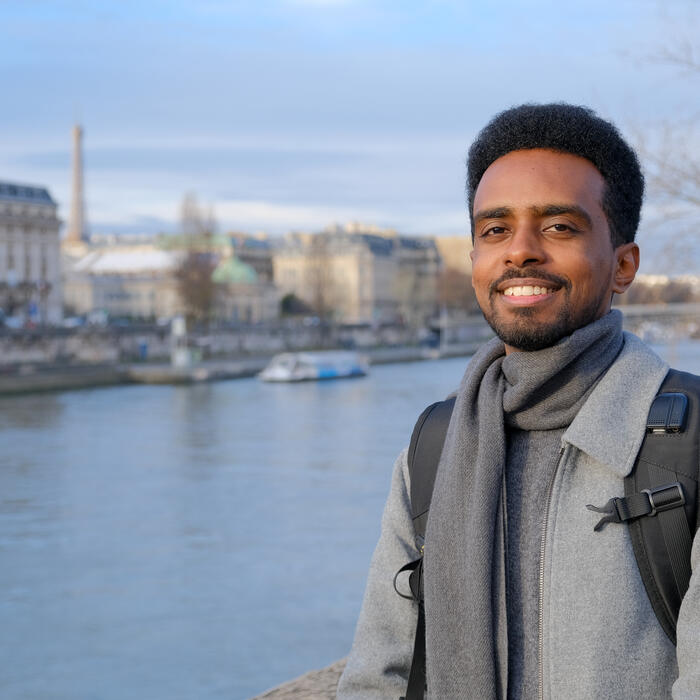 Person standing by a river in a city, wearing a grey coat and scarf with a backpack, with historic buildings and a bridge blurred in the background.