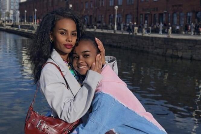 Two people by a waterfront, standing close together with buildings and water in the background.